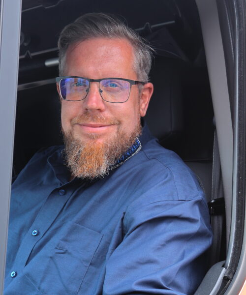 Simon Vieten sitzt hinter dem Steuer eines Kleintransporters und blickt, freundlich lächelnd, aus dem Seitenfenster des Kleintransporters heraus.